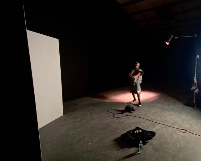 A person stands in a dimly lit studio with a violin, near a white panel and some lighting equipment.