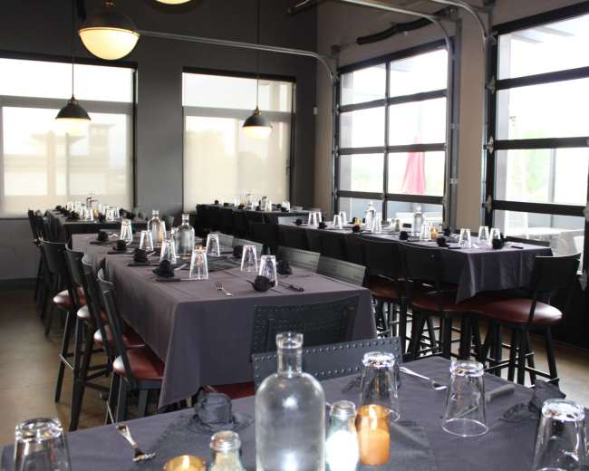 A spacious dining room features several long tables set with dark tablecloths, glassware, and black napkins, under large windows with a modern industrial design.