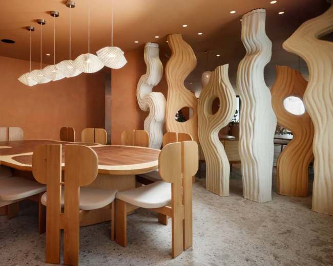 The image shows a modern dining room with a round wooden table surrounded by unique wooden chairs, illuminated by pendant lights, and accented by sculptural wall features.