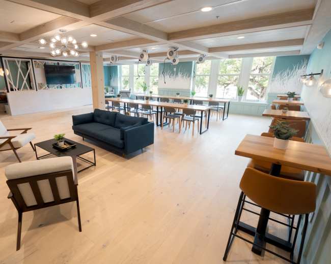 The image features a spacious, modern lounge area with a mix of seating arrangements, including a dark sofa, wooden tables, and chairs, alongside large windows that allow natural light to fill the room.