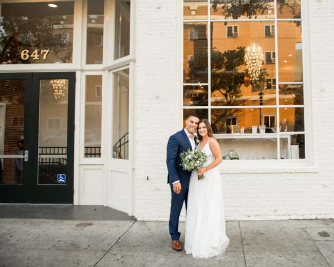 A couple in formal attire stands outside a white brick building with large windows, holding a bouquet of flowers.