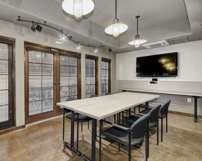 A meeting room features multiple tables and chairs, large windows with blinds, and a wall-mounted television.