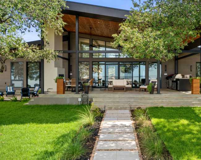 A modern house with large windows and a covered patio is situated on a grassy lawn, with a stone pathway leading to the entrance.