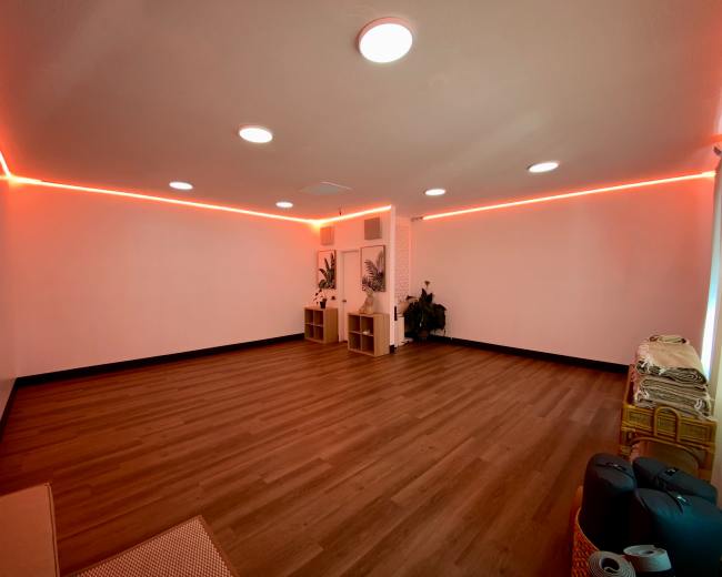 The image shows a spacious room with wooden flooring and red LED strip lighting along the walls, featuring two storage units and a few yoga mats rolled up in the corner.