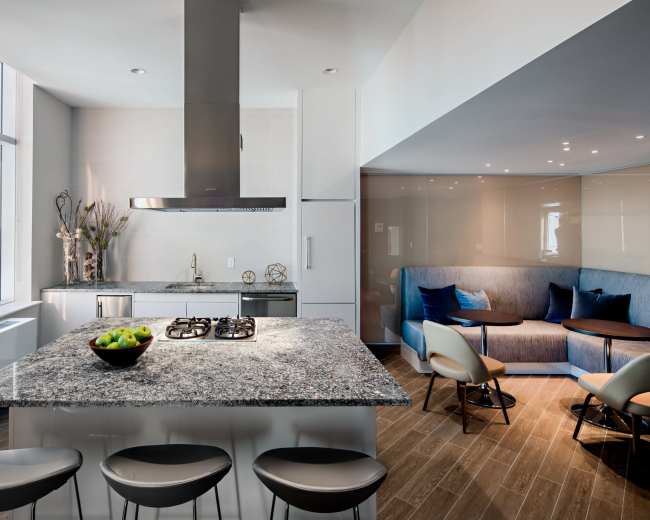 The image shows a modern kitchen featuring a large granite island with a gas cooktop, sleek cabinetry, and a seating area with curved chairs and a sofa, all illuminated by natural light from large windows.