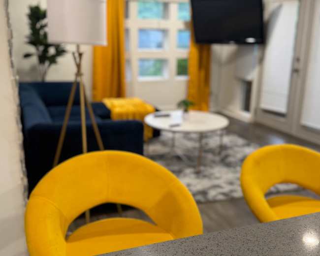 A modern living room features a blue sofa, a round coffee table, and yellow curtains, with two yellow chairs in the foreground.