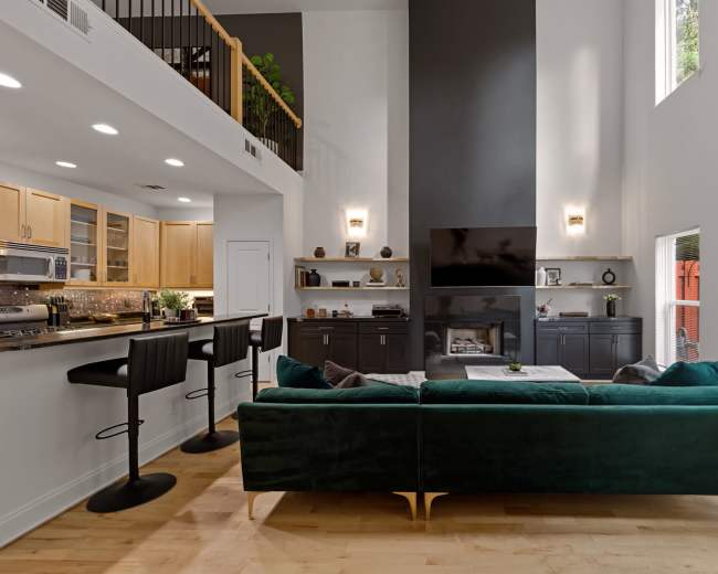 The image shows a modern living room with a green velvet sofa facing a sleek fireplace and a wall-mounted television, adjacent to a kitchen with two bar stools at the counter.