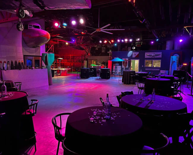 The image shows a dimly lit event space with round tables set for dining, colorful lights illuminating the area, and a vintage airplane hanging from the ceiling.