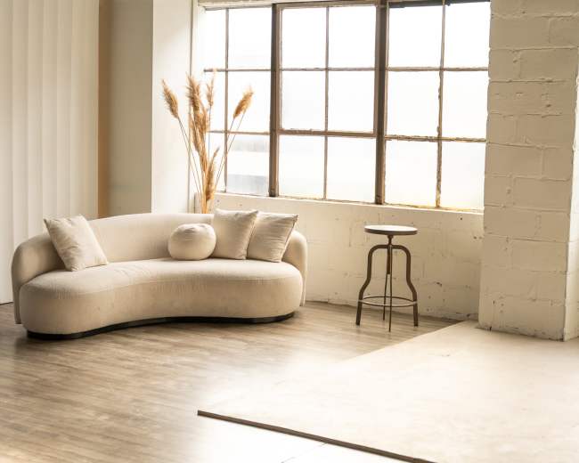 The image displays a modern, curved beige sofa next to a circular wooden table in a bright, minimalist room with large windows.