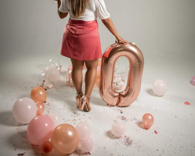 A woman stands beside large rose gold "30" balloons, surrounded by pastel balloons on the floor.