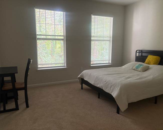 A sparsely furnished bedroom features a bed with a white comforter and a small desk with a chair near two windows.