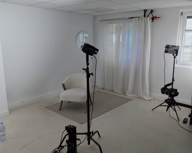 The image shows a minimalist studio space with a white chair, a curtain, a mirror on the wall, and two lighting stands positioned on either side.
