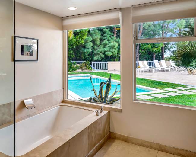 A modern bathroom features a bathtub beside a large window that opens to a backyard pool area with lounge chairs.