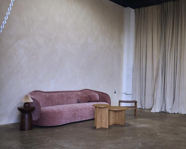 The image features a minimalist interior with a pink sofa, two wooden side tables, and a curtain backdrop.