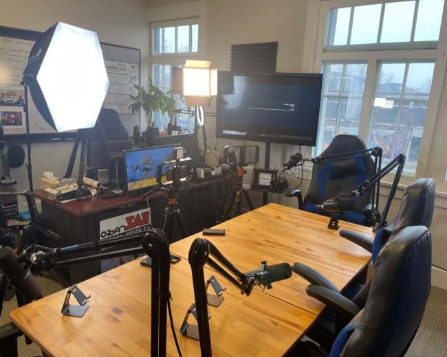 The image shows a room set up for a recording, featuring a wooden table with several microphone stands, office chairs, and lighting equipment.