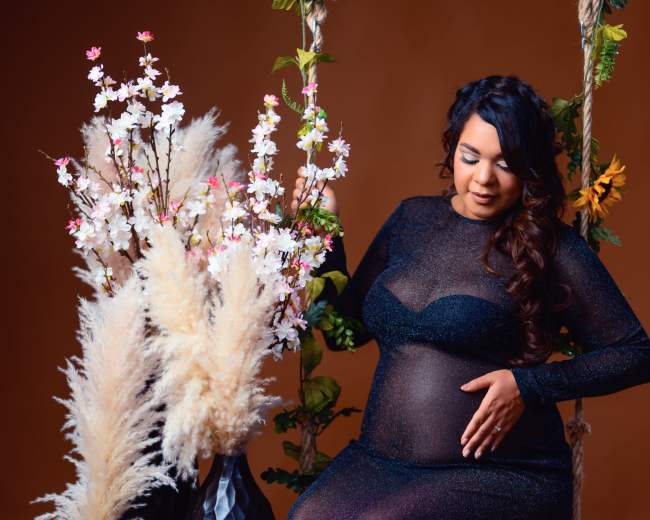 A pregnant woman in a black, sheer dress gently touches her belly while sitting on a swing beside decorative vases filled with flowers and pampas grass.