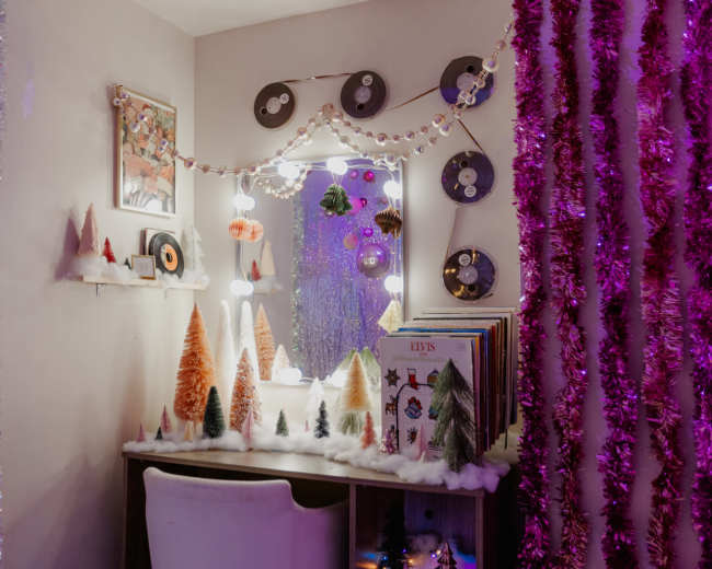 A small dressing table decorated with miniature trees and festive ornaments is illuminated by a mirror and surrounded by colorful tinsel and vintage records.