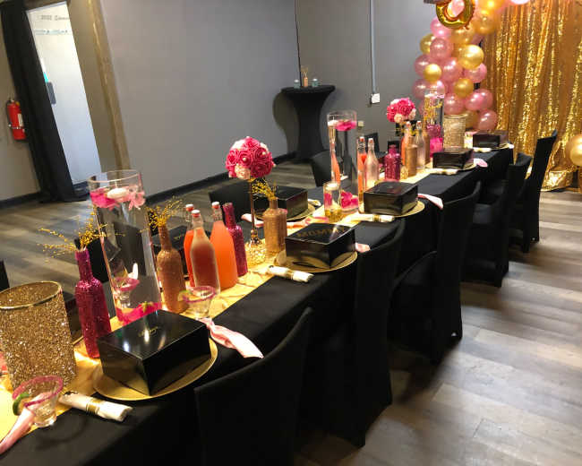 A decorated banquet table with black chairs, adorned with colorful bottles, floral centerpieces, and a gold table runner.
