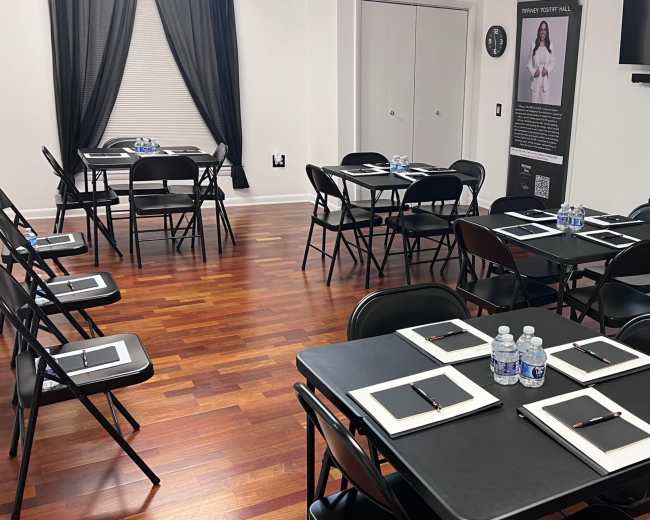 The image shows a room set up with multiple black tables and chairs, each equipped with notebooks and bottled water, in a space with hardwood flooring and a television on the wall.