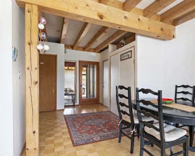 A dining area with a round black table and six chairs, set against a background of wooden beams and an adjoining hallway.