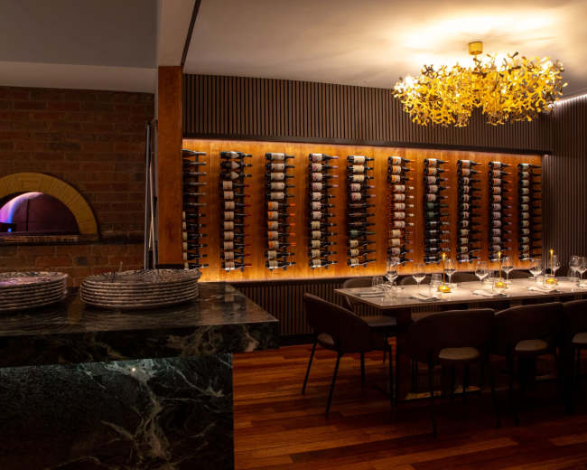 The image shows a modern dining area featuring a wine wall, a large wooden table set with dinnerware, and a stone pizza oven in the background.