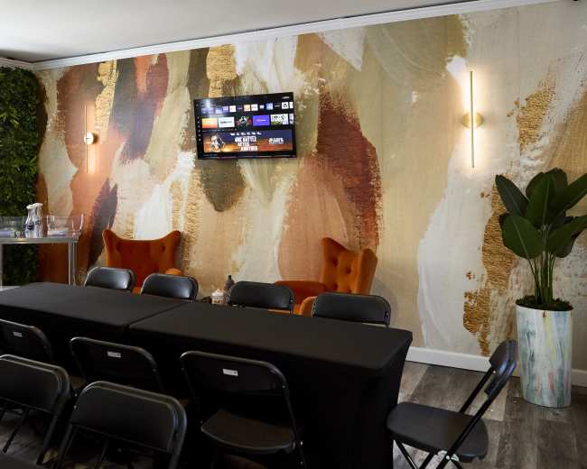 The image shows a modern conference room with a large patterned wall, black tables arranged in rows, and orange accent chairs.