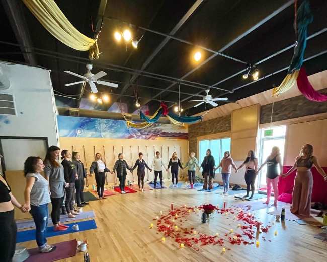 A group of people stands in a circle, holding hands around a flower and candle arrangement on the wooden floor of a spacious room with colorful decorations hanging from the ceiling.