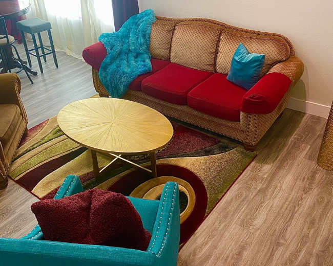 The image shows a living room with a red and beige sofa, a round gold coffee table, a turquoise armchair, and a floral painting on the wall.