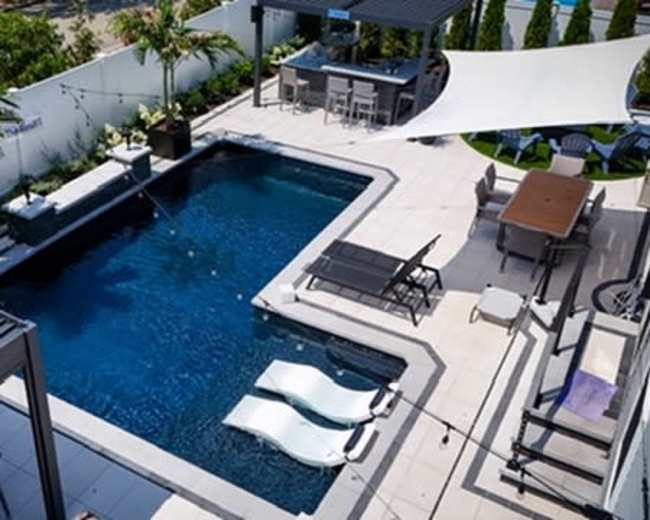 The image shows a modern backyard featuring a rectangular swimming pool surrounded by lounge chairs, a dining area, and shaded structures.