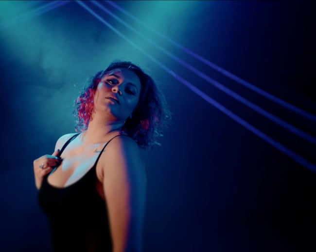 A person with curly hair poses under blue and purple lighting in a smoky environment.