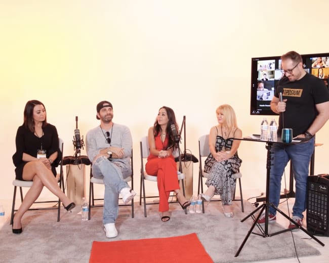 A panel of five individuals is seated on chairs in front of a screen displaying a video call, with a small table and microphones in the foreground.