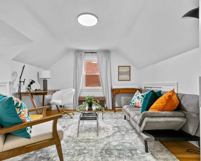 A cozy living space with a sloped ceiling, featuring two armchairs and a sofa arranged around a central coffee table, a desk in one corner, and a window with light curtains.