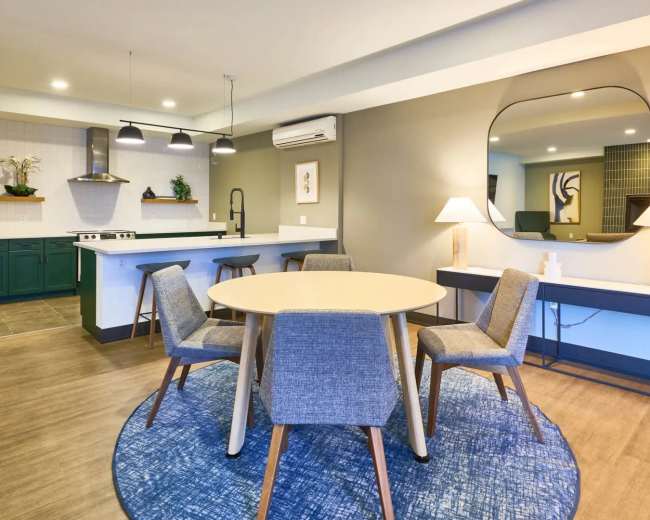 The image shows a modern kitchen and dining area featuring a round table with four chairs, a large mirror on the wall, and a kitchenette with green cabinets and a sleek countertop.