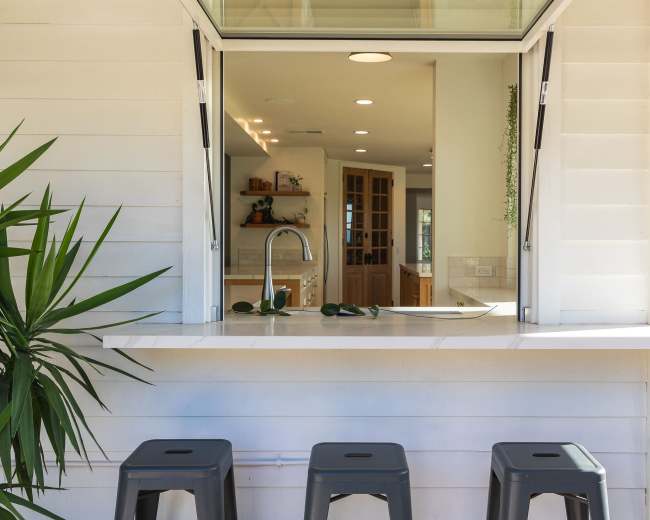 A wide opening connects an outdoor bar with three gray stools to an interior kitchen area featuring wooden shelves and cabinets.