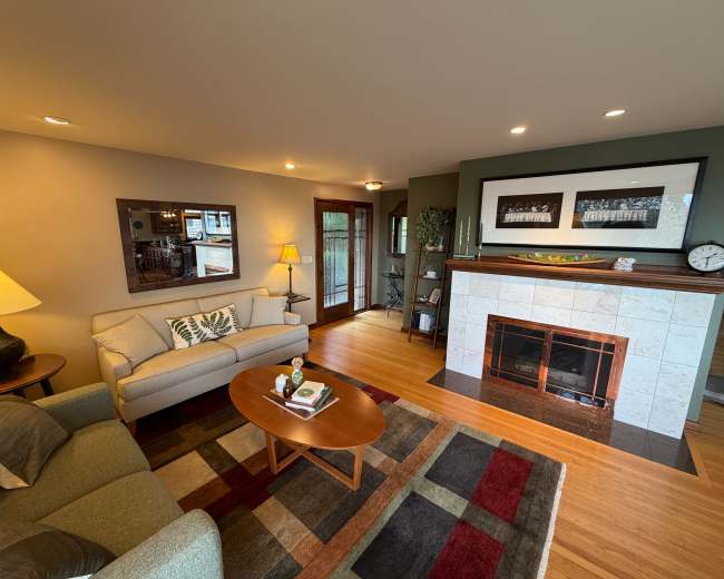 A cozy living room features a sofa, an oval coffee table, a fireplace with decorative tiles, and hardwood flooring.