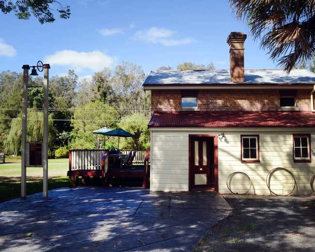 The image shows a two-story brick and wooden house with a red roof, a small deck, and a seated person under an umbrella in a green outdoor area.