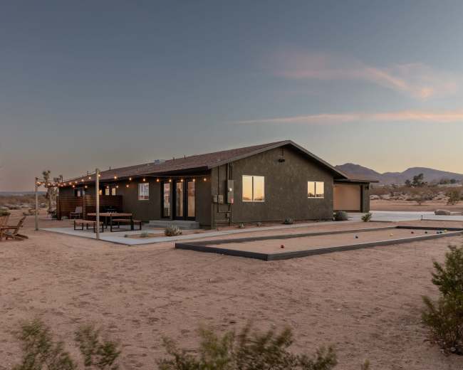A modern single-story house sits in a desert setting, with a fire pit and a bocce ball court in the foreground.