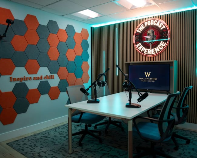 The image shows a podcast studio featuring a table with microphones, a screen displaying text, and colorful wall decor made of hexagonal panels.
