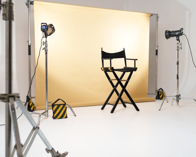 A photography studio features a folding director's chair in front of a yellow backdrop, surrounded by lighting equipment on stands.