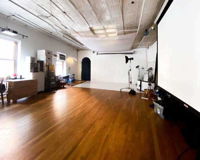 The image shows a spacious, empty studio with wooden flooring, large windows, and a blank white wall at the front, along with some equipment and storage in the back.