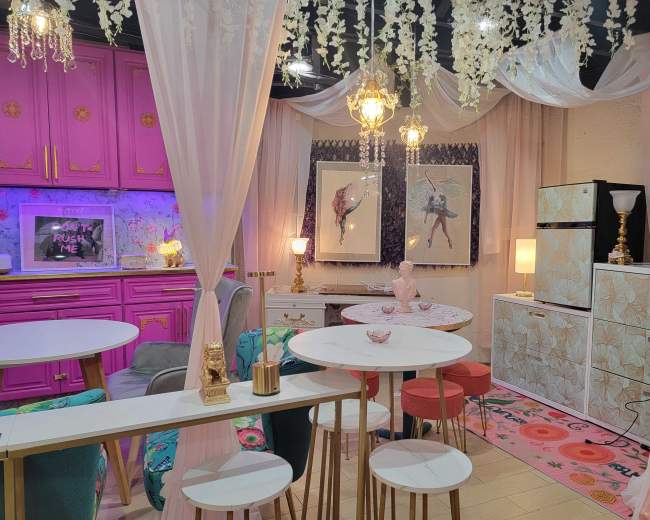 The image shows a vibrant, stylish interior featuring pink cabinetry, various decorative furnishings, and multiple light fixtures hanging from the ceiling.