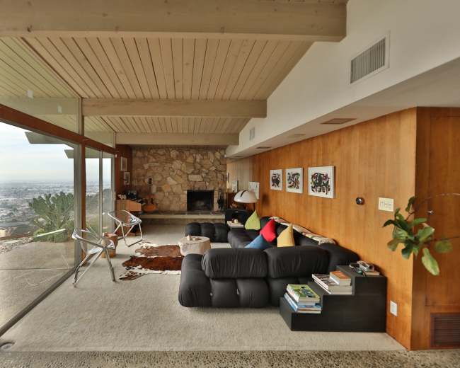 The image shows a modern living room with large glass windows, a stone fireplace, and a mix of furniture including a black sofa, a chair, and a decorative area rug.