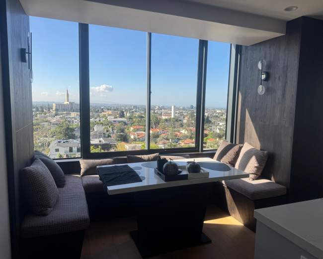 A modern dining nook with a table and cushioned seating offers a view of a city skyline through large windows.