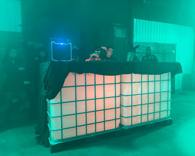 A DJ booth made of stacked white containers is illuminated with green and orange lighting in a warehouse setting.
