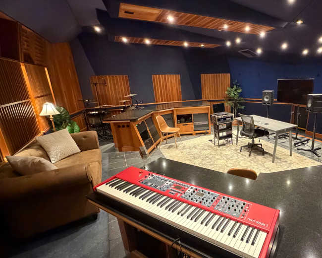 A music recording studio features a red keyboard on a countertop, surrounded by instruments, desks, and studio equipment in a spacious room with acoustic panels.