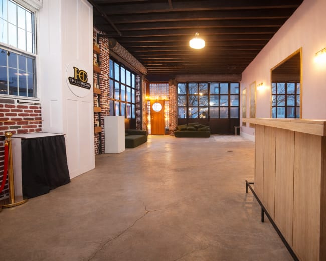 The image shows a spacious, modern interior with exposed brick walls, large windows, and a wooden reception desk.
