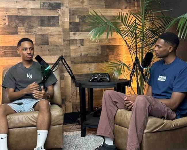 Two young men are seated on brown chairs in a recording studio with a wooden wall and green plants, engaged in conversation while microphones are positioned nearby.