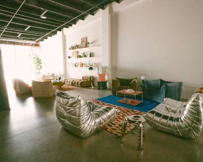 The image shows a spacious modern living area with several unique seating arrangements, a blue rug, and shelves displaying decor items.