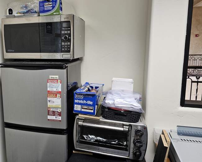 A microwave is placed on top of a compact refrigerator, with kitchen items stacked nearby, including a toaster oven and some bags.