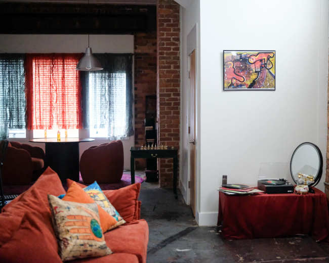 The image shows a cozy living space with a brick wall, a red sofa adorned with colorful pillows, and a record player on a table next to a framed artwork on the white wall.